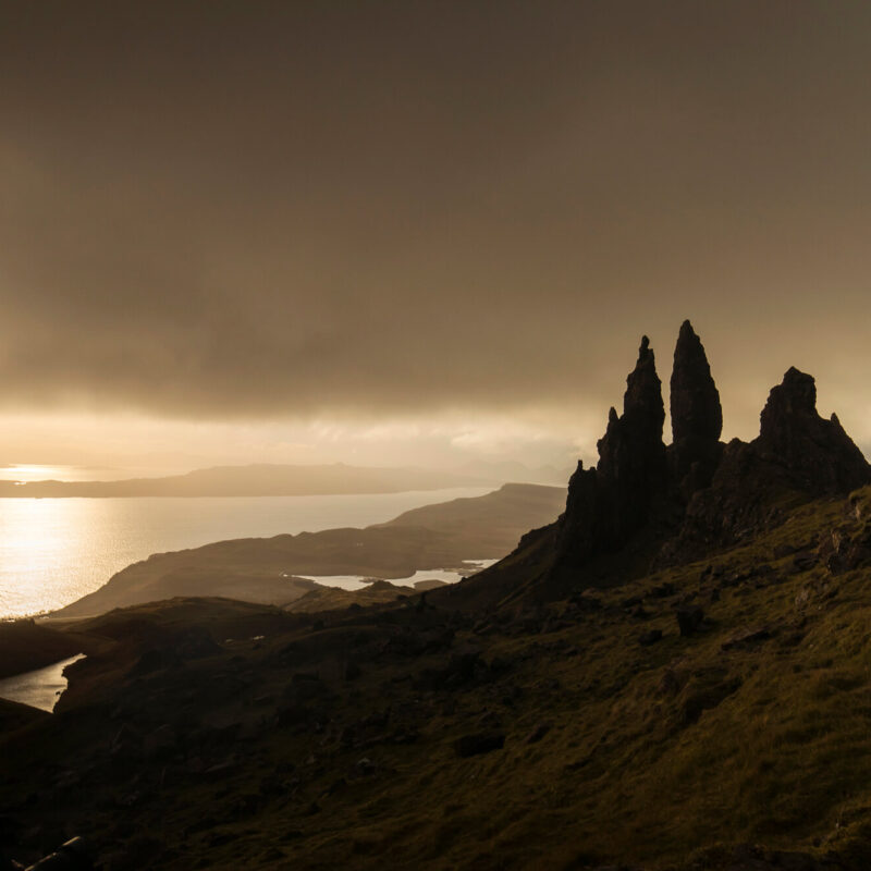 Dramatic landscape view of the Old Man of Storr at sunrise on the Isle of Skye, with warm light over the coastline and rocky pinnacles.