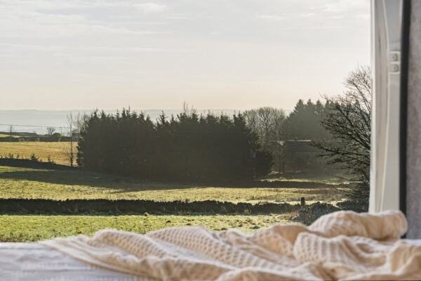 Campervan rear doors open to a countryside view with soft bedding and natural light inside.