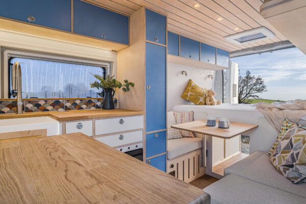 Wide view of campervan kitchen and seating area with blue cabinetry and timber ceiling.