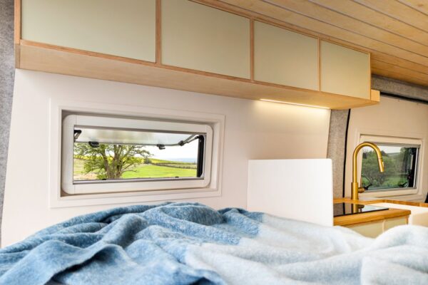 Campervan fixed bed with overhead lockers and countryside views through opening window